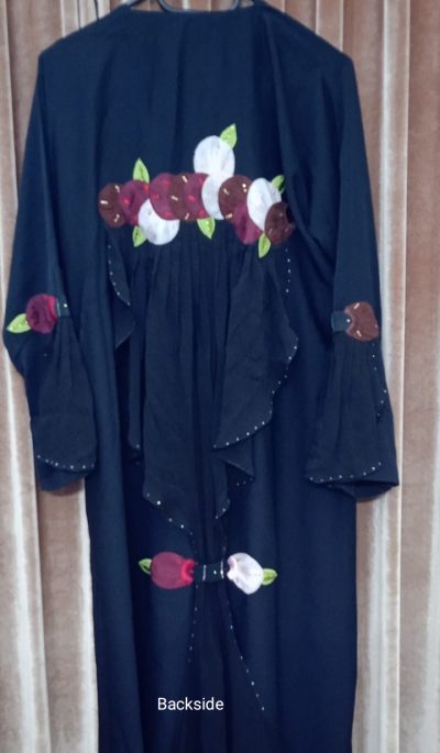 The backside of a black abaya showing a large horizontal floral applique design across the upper back and ruffled fabric panels.