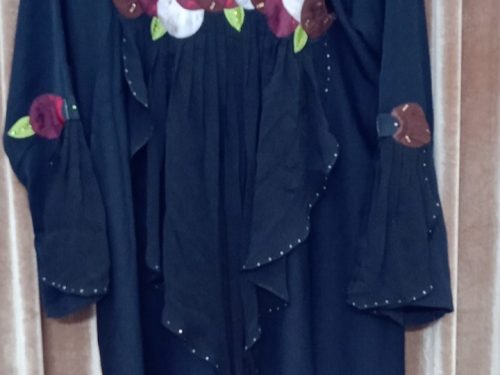 The backside of a black abaya showing a large horizontal floral applique design across the upper back and ruffled fabric panels.