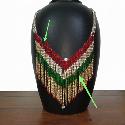 A V-shaped fringe necklace featuring red, white, green, and gold rhinestones on a black display bust. Green arrows point to missing beads and broken strands in the fringe.
