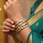 A pair of thick, two-tone bangles featuring a braided or wheat-link design in gold and frosted silver, accented with small embedded crystals. The bangles are worn on a wrist against a green and gold saree.