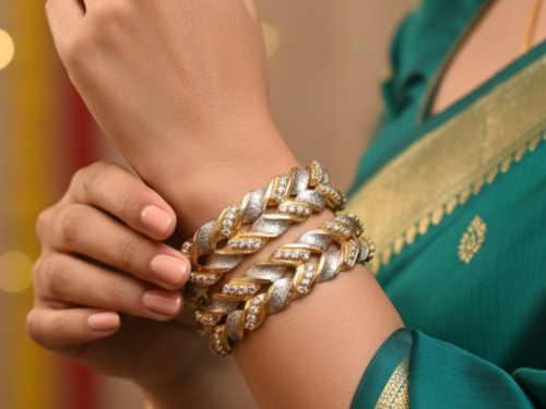 A pair of thick, two-tone bangles featuring a braided or wheat-link design in gold and frosted silver, accented with small embedded crystals. The bangles are worn on a wrist against a green and gold saree.