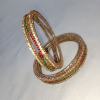 A pair of ornate, gold-toned bangles featuring a textured, lattice-like design. Each bangle is detailed with a circular row of small white crystals, accented by alternating sections of vibrant red and green stones.