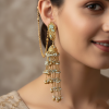 A close-up of a woman smiling, wearing a large, intricate gold Jhumka earring. The earring is decorated with mint green and white beads. A delicate gold chain (ear string) wraps from the earring stud up to the top of her ear, tucked into her hair.