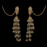 A pair of gold-toned Jhumka earrings displayed against a solid black background. The earrings feature sea-green beaded studs, a bell-shaped centerpiece, and multiple cascading chains adorned with tiny white beads and gold droplets. Long "Sahara" ear chains extend from the studs.