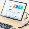​A tablet on a desk stand displaying a cryptocurrency financial dashboard, connected to a multi-port USB-C docking station. The setup includes a mechanical keyboard, a mouse, and several peripherals plugged into the silver hub, emphasizing a portable "desktop" workspace on a light wood surface.