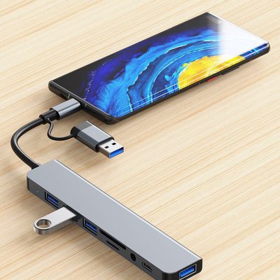 A silver multi-port USB-C hub adapter connected to a smartphone on a light wood surface. The hub features a 2-in-1 connector (USB-C and USB-A), several USB ports, an SD card slot, and a headphone jack. A silver USB flash drive is plugged into one of the side ports.