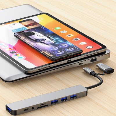A slim, space-gray aluminum USB hub featuring a dual-head connector (USB-C and USB-A) sitting on a wooden desk. The hub includes four USB 3.0 ports, a USB-C port, a 3.5mm headphone jack, and SD/TF card slots. In the background, a laptop, tablet, and smartphone are stacked, showcasing the hub's compatibility across multiple devices.