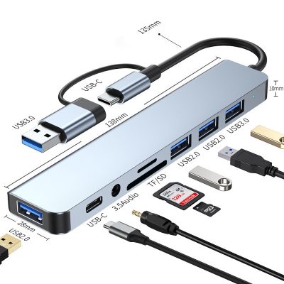 A sleek grey 7-in-1 USB-C docking station featuring a dual USB-C and USB-A connector cable, displayed on a wooden surface next to a stacked laptop, tablet, and smartphone.